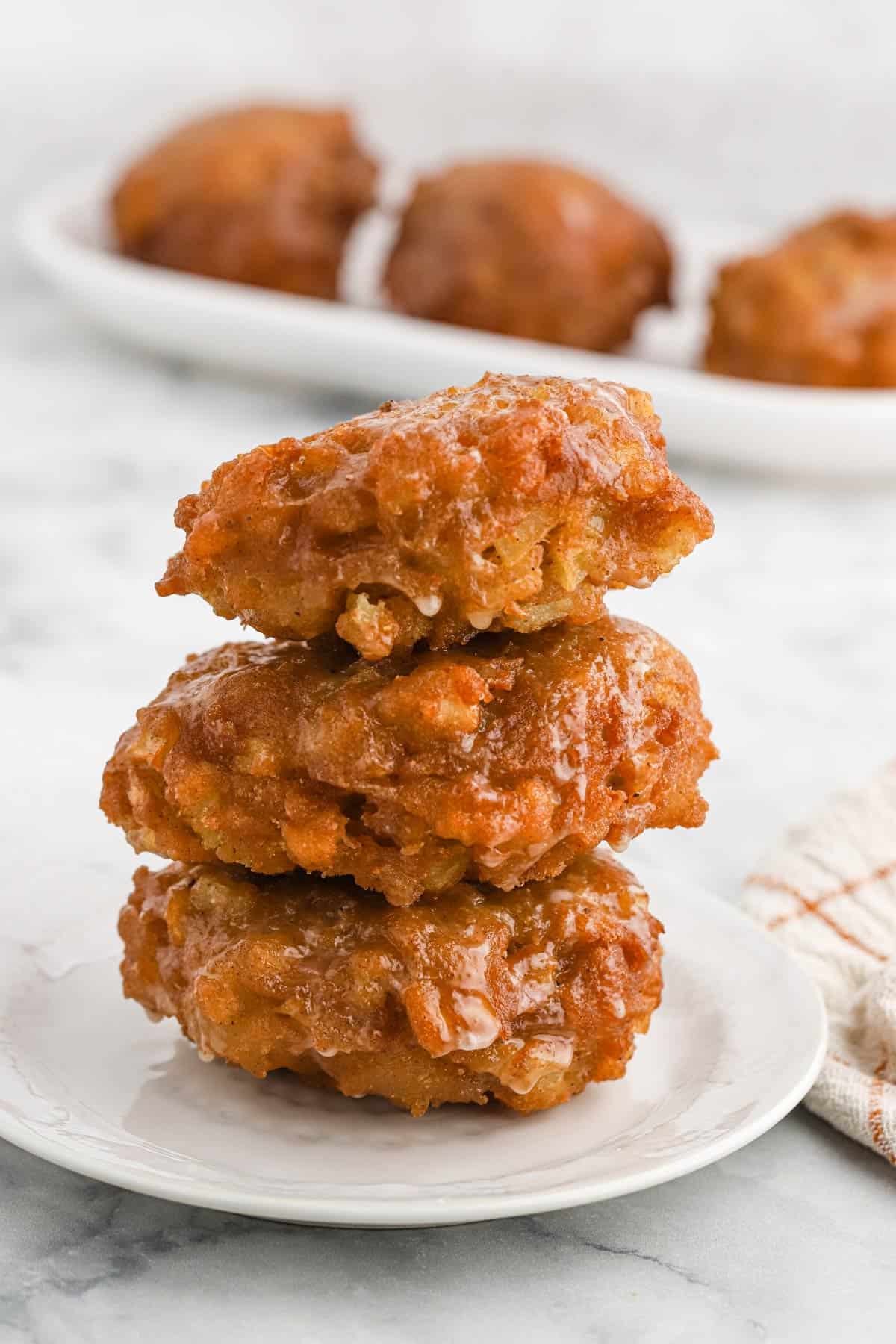 A stack of apple fritters on a white plate with more on a tray in the background.