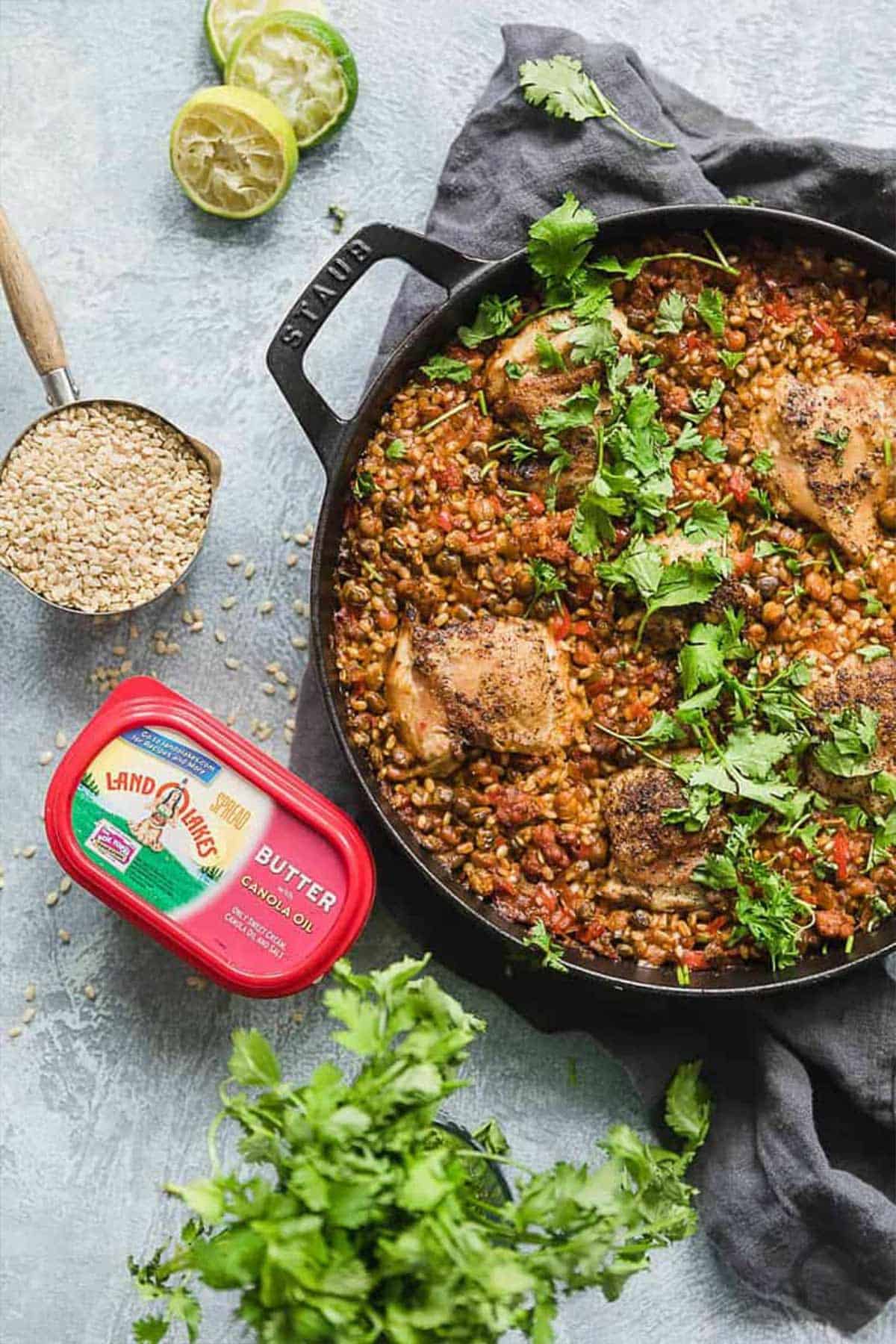 A dish of arroz con pollo on the table with a measuring cup of brown rice, carton of butter, and squeezed limes on the table.