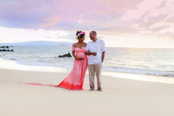 Jocelyn wearing a flower head wrap with her husband stand on the sands of a beach in Maui, Hawaii