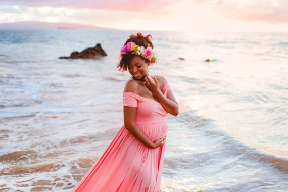 Jocelyn smiling on a beach in Maui dressed in a pink maternity dress at 30 weeks pregnant