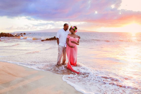 Jocelyn and her husband standing in the ocean water on a beach in Hawaii with the sun in the background setting