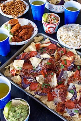 Game Day Pizza Nachos served on a metal baking tray along with pretzels, hot wings, popcorn and blue plastic cups full of a beverage