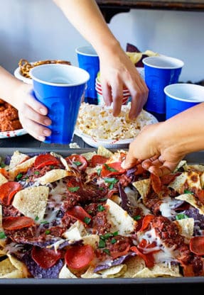 Three hands grabbing at Game Day Pizza Nachos and popcorn with wings and blue plastic cups in the background