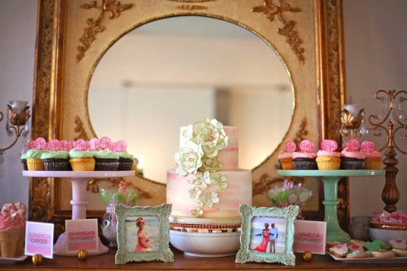 Dessert table full of cupcakes, cookies, photos and a large white and pink cake in the middle