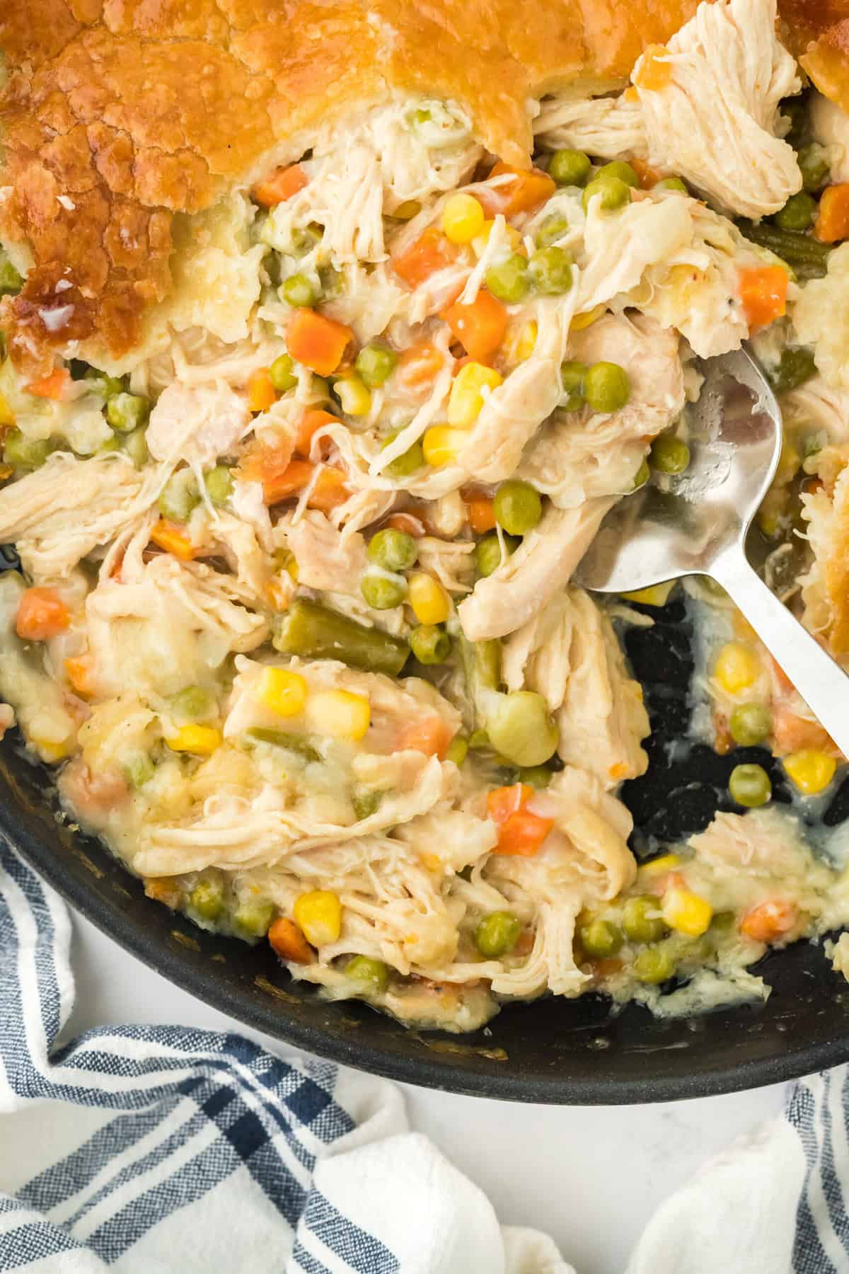 An overhead of the best chicken pot pie recipe with a spoon digging into the skillet on a white background with a striped napkin