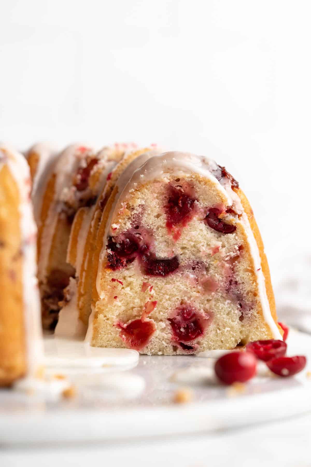 Cranberry Orange Cake with two slices cut out. Those slices are sitting on their own plates in front of the cake with a bowl of cranberries and oranges surrounding them. 