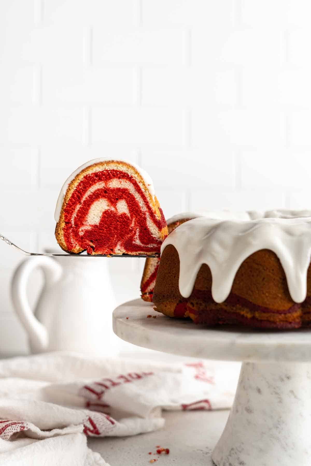Red Velvet Marble Cake is sliced on white cake stand with white background with a spatula lifting a piece of cake up to show the swirl pattern.
