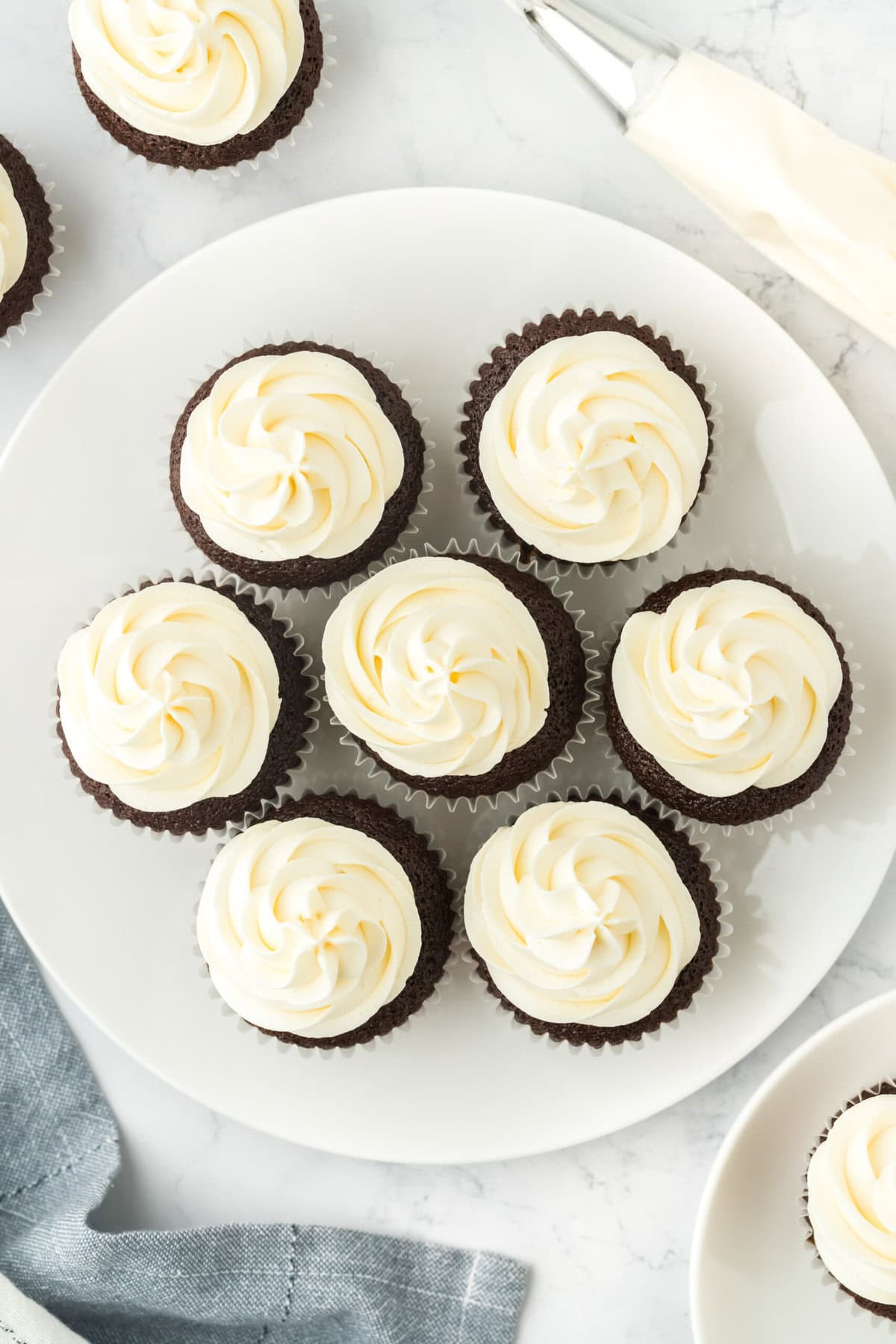 An overhead of easy chocolate cupcakes on a white plate with white frosting and a piping bag filled with frosting nearby
