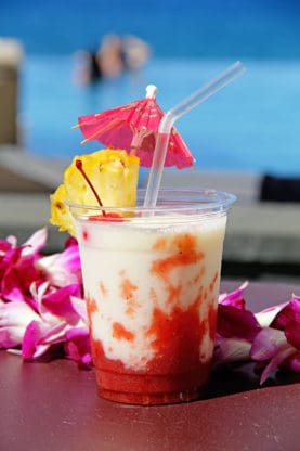 Close up of a plastic cup full of a lava flow drink with a cherry, drink umbrella and pineapple wedge in it and a lei next to it