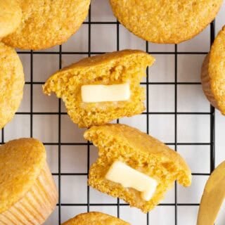 A split cornbread muffin with two pats of butter melting inside, surrounded by more muffins on a cooling rack