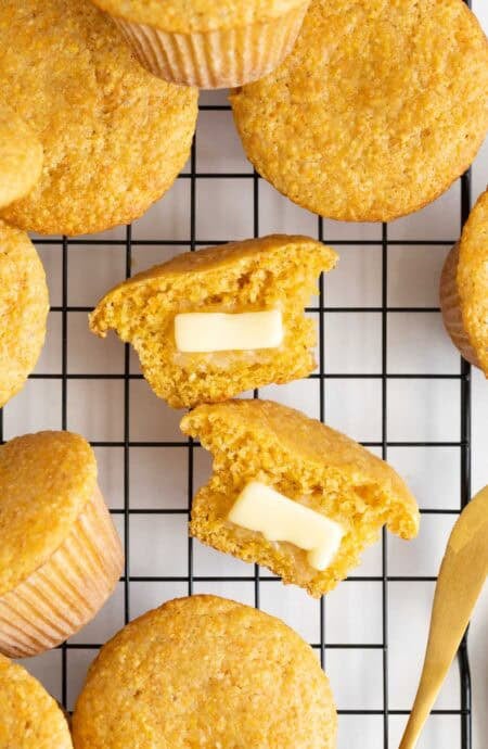 A split cornbread muffin with two pats of butter melting inside, surrounded by more muffins on a cooling rack