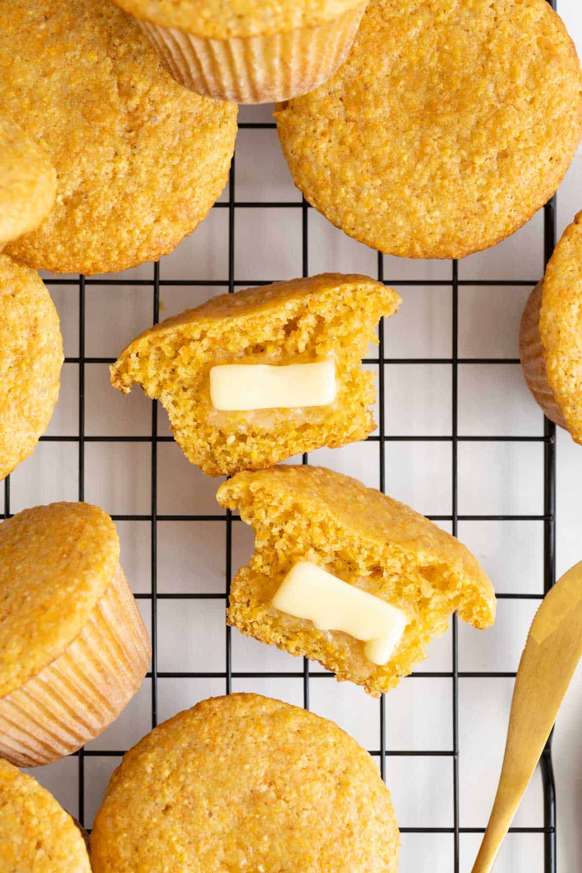 A split cornbread muffin with two pats of butter melting inside, surrounded by more muffins on a cooling rack