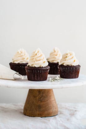 Easy Chocolate Cupcakes sitting on a white cake plate next to a frosting piping tool.