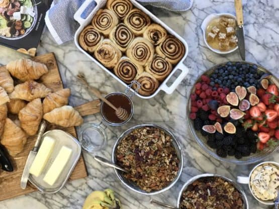 Overhead shot of foods including a bowl of a variety of fruits, croissants and butter, cinnamon rolls and other items on a marble background