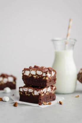 Two Mississippi Mud Brownies stacked on each other with marshmallows and nuts next to them and a glass of milk with a straw in it in the background
