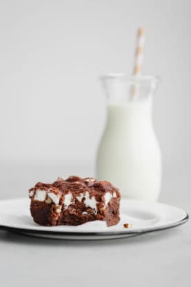 A half eaten Mississippi Mud Brownie served on a white, round plate with a glass of milk with a straw in the background