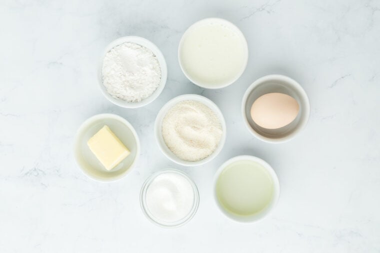 Flour, sugar, butter, egg, cornmeal in white bowls on white countertop