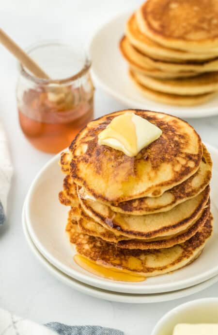 Hoe cakes in a giant stack on a white plate with butter on them and honey in the background