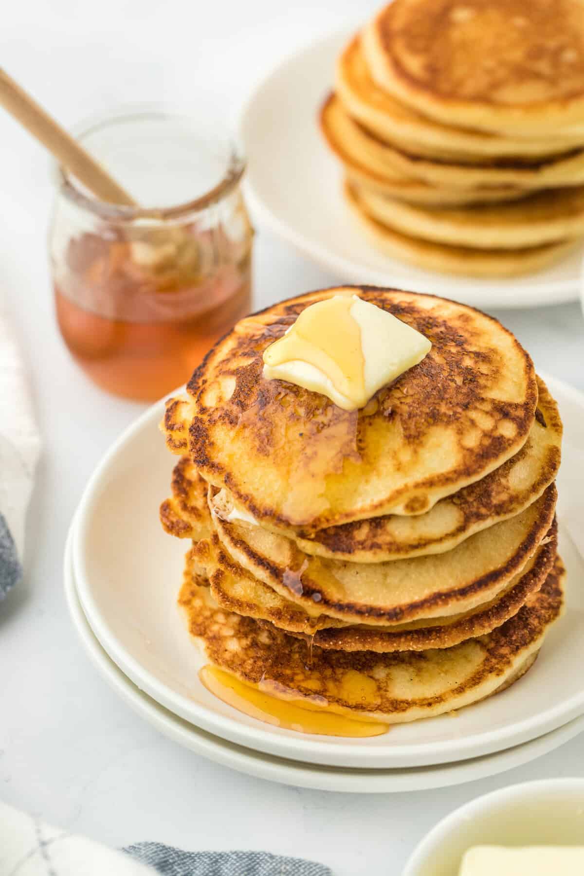 Hoe cakes in a giant stack on a white plate with butter on them and honey in the background