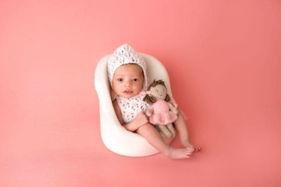 Harmony wearing a white knit outfit and hat sitting on a small white chair with a baby doll
