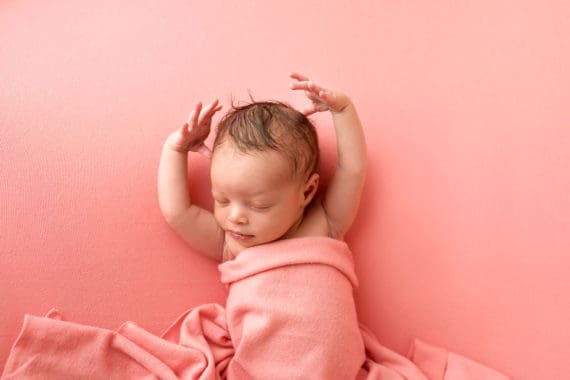 A sleeping Harmony holding her arms up and covered in a pink blanket on a pink background