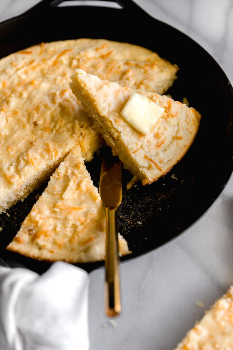 Close up of Mexican cornbread recipe slice with butter against full cast iron skillet of cornbread ready to serve