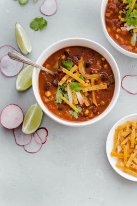 Overhead shot of a cup of Chipotle Black Bean Tortilla Soup with a spoon in it, another bowl of soup behind it, a small plate of tortilla chip strips and slices of radish and lime surrounding them