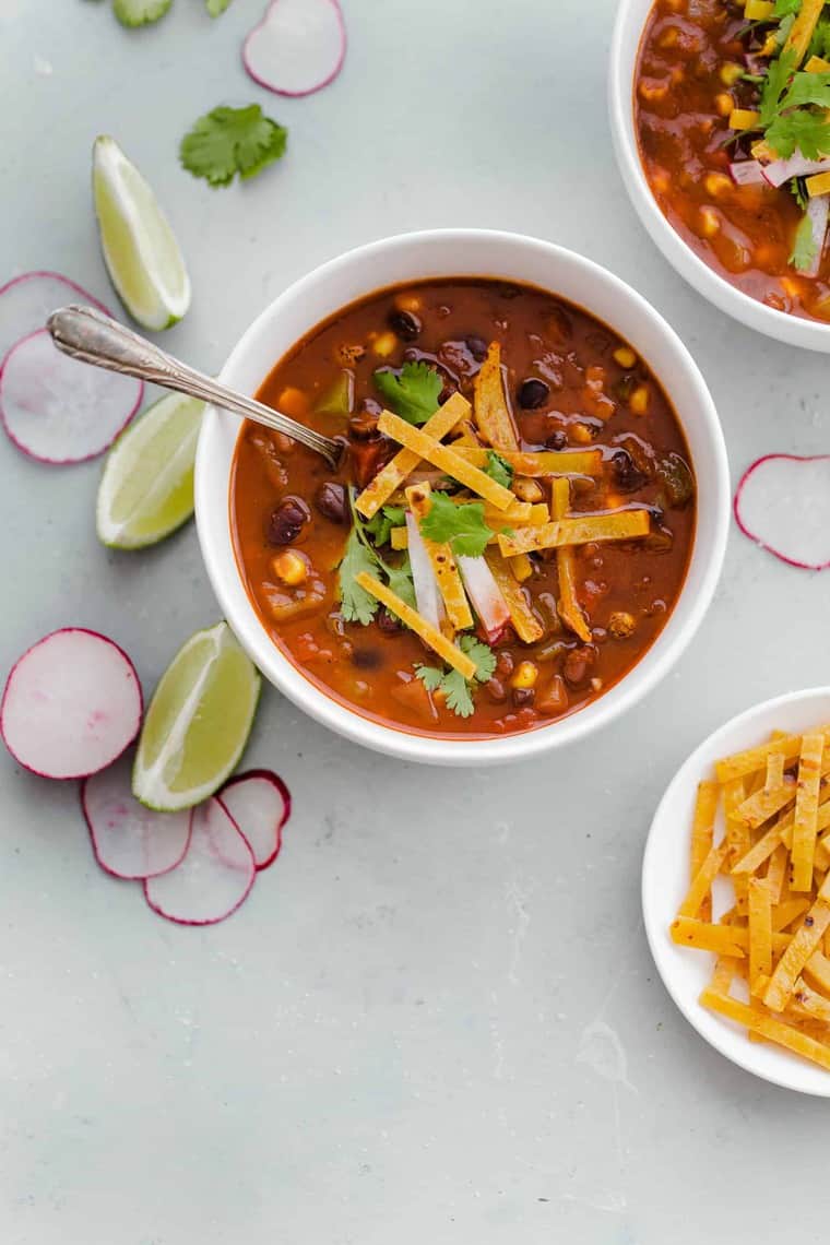 Overhead shot of a cup of Chipotle Black Bean Vegetarian Tortilla Soup with a spoon in it, another bowl of soup behind it, a small plate of tortilla chip strips and slices of radish and lime surrounding them