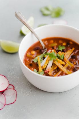 A cup of Chipotle Black Bean Tortilla Soup with a spoon in it and pieces of radish and lime next to it