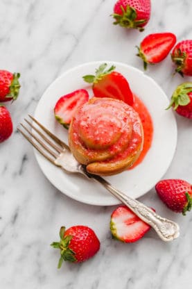 Overhead shot of one Strawberry Butter Rose Bun served on a white plate with a fork and topped with strawberry glaze surrounded by fresh strawberries