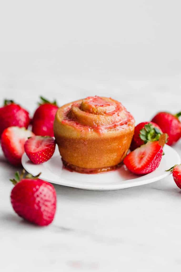 One Strawberry Butter Rose Bun served on a white plate and topped with strawberry glaze surrounded by fresh strawberries