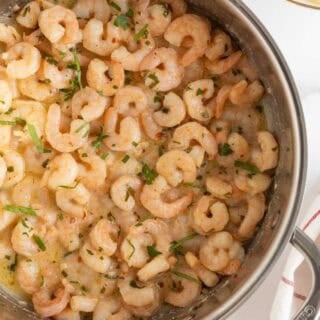 Overhead shot of a stainless steel pan filled with easy shrimp scampi, garnished with chopped fresh parsley, on a white marble surface with a blurred bowl of pasta in the background