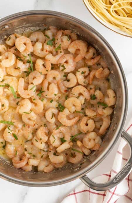 Overhead shot of a stainless steel pan filled with easy shrimp scampi, garnished with chopped fresh parsley, on a white marble surface with a blurred bowl of pasta in the background