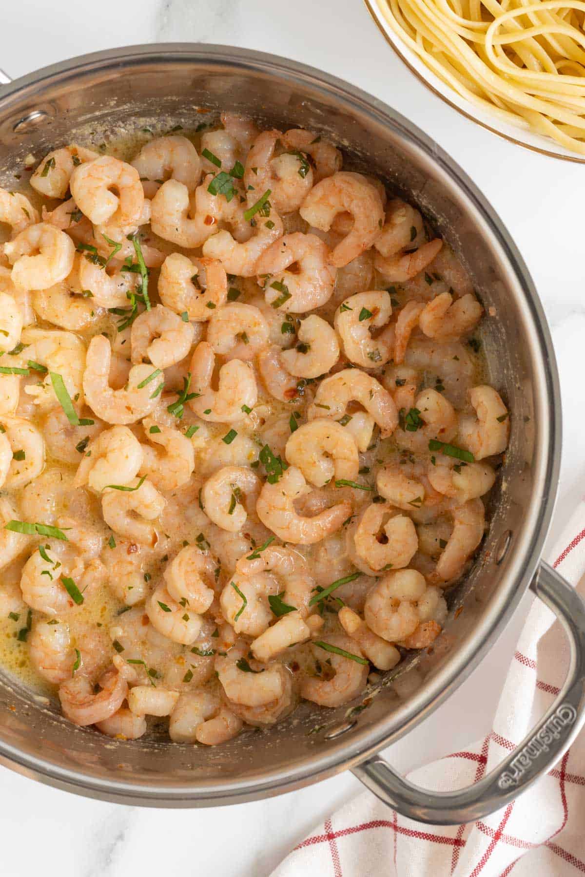 Overhead shot of a stainless steel pan filled with easy shrimp scampi, garnished with chopped fresh parsley, on a white marble surface with a blurred bowl of pasta in the background