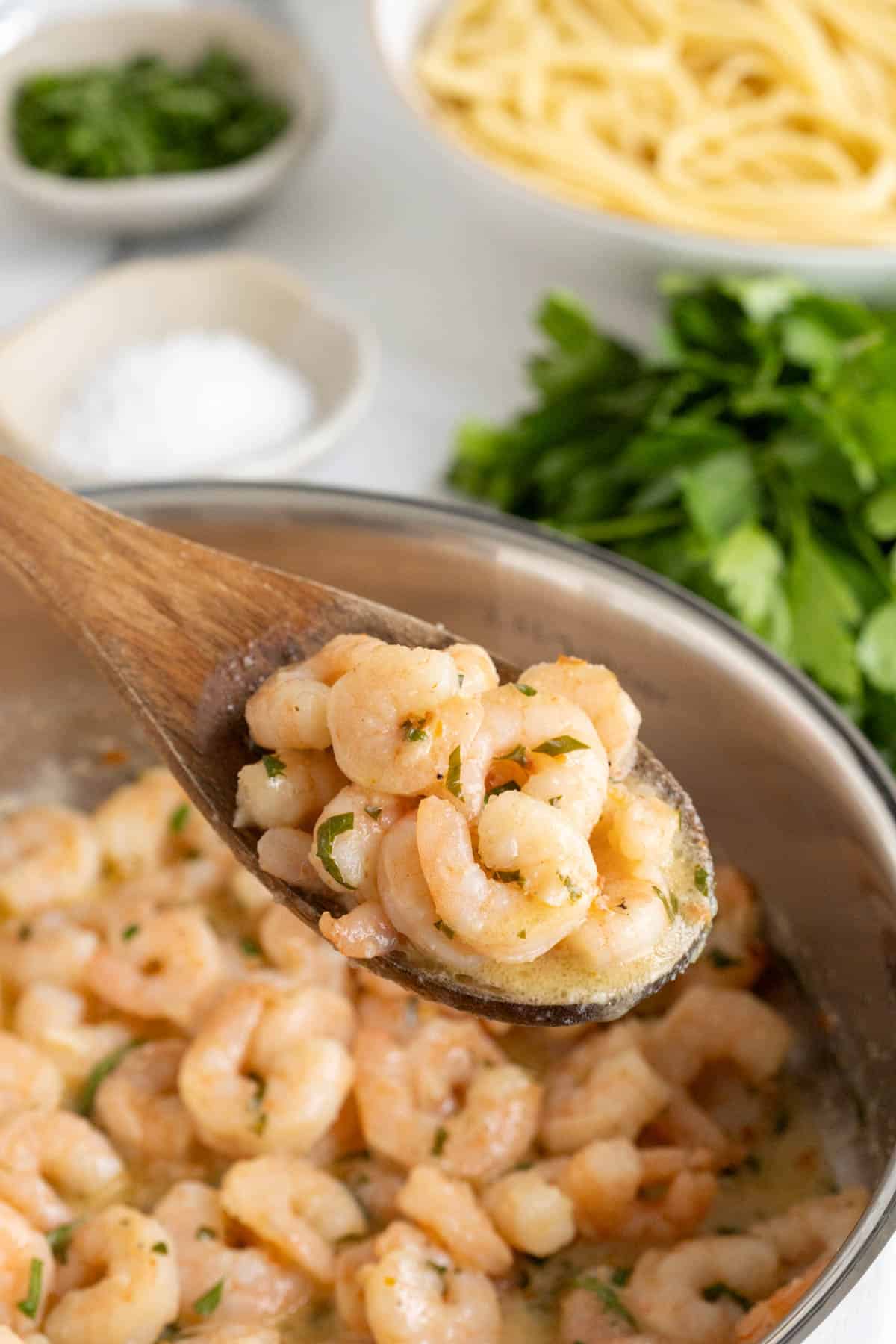 A wooden spoon holding several cooked shrimp in a creamy sauce, lifted from a pan of more easy shrimp scampi, with blurred salt, parsley, and pasta in the background