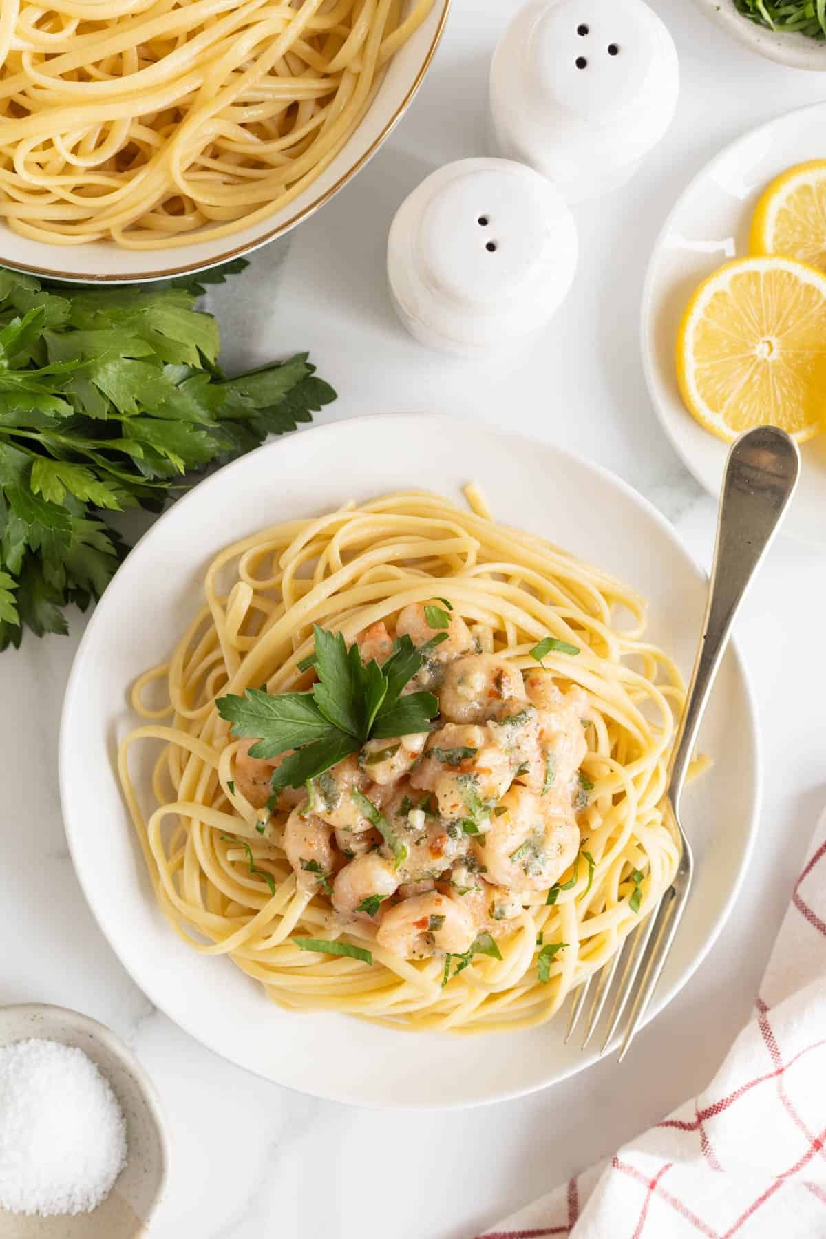 Overhead shot of a white plate with spaghetti topped with a generous serving of shrimp scampi and a sprig of parsley, with lemons, salt, and more pasta blurred in the background