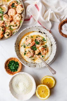 Shrimp Scampi and grits in a bowl surrounded by lemons, uncooked grits and oregano.