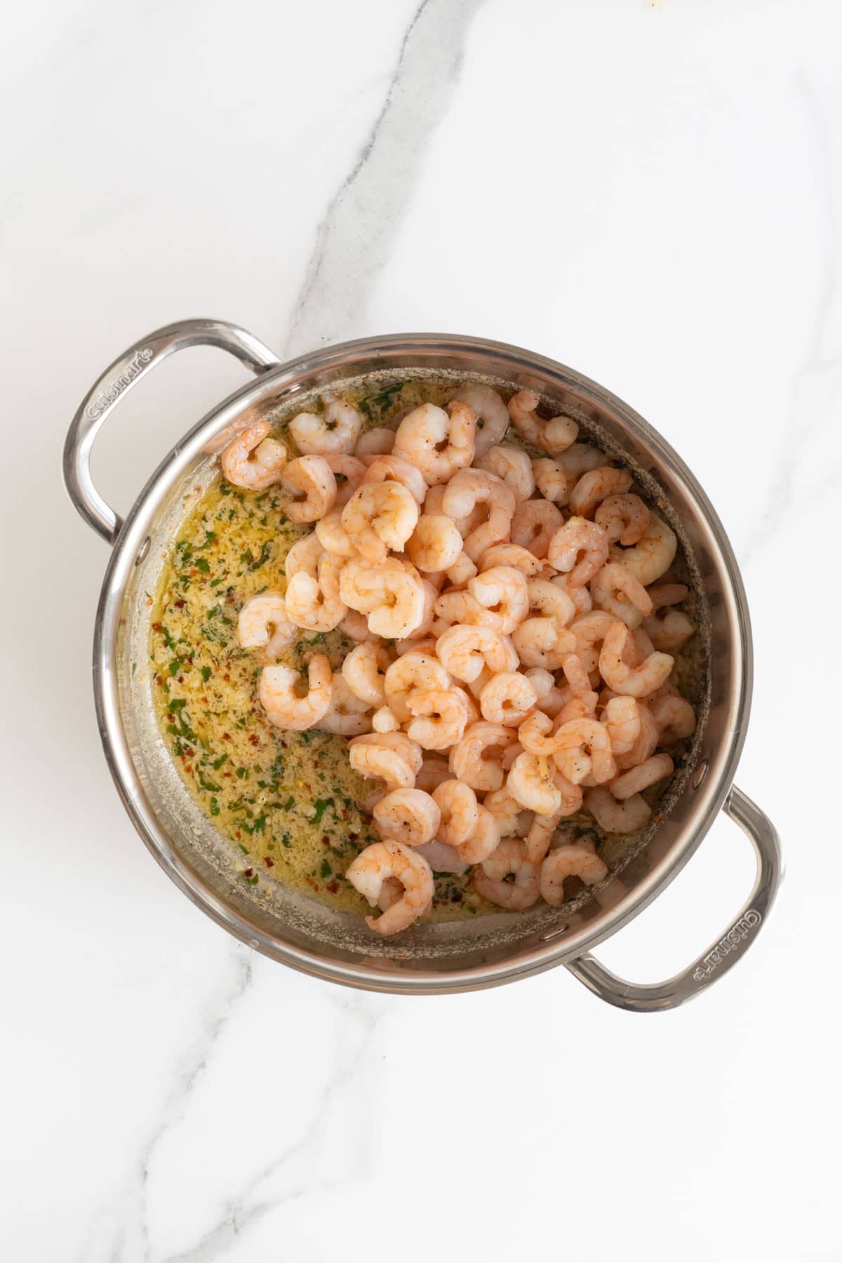 Overhead shot of a stainless steel pan containing cooked shrimp and a liquid base for scampi, with chopped parsley, red pepper flakes, and minced garlic
