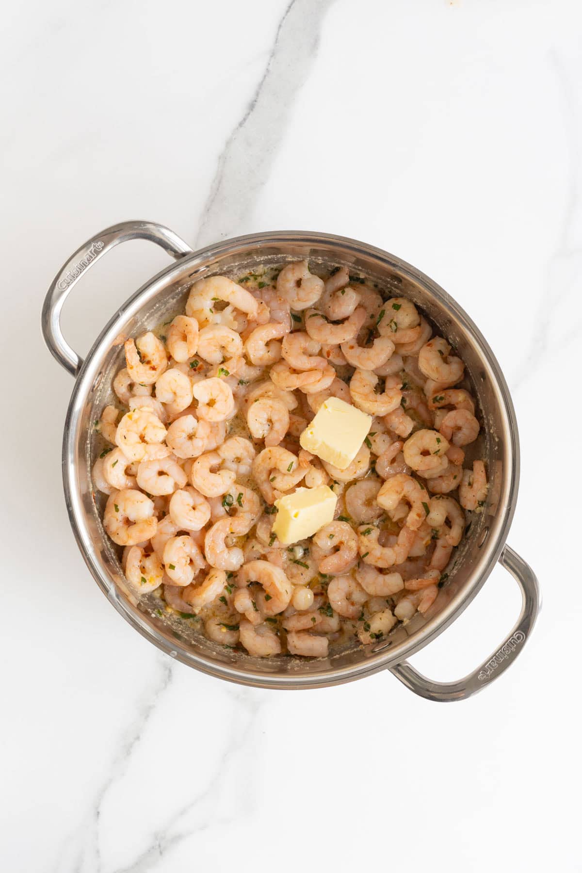 Overhead shot of a stainless steel pan filled with cooked shrimp in sauce, with two pats of butter added on top, ready to melt