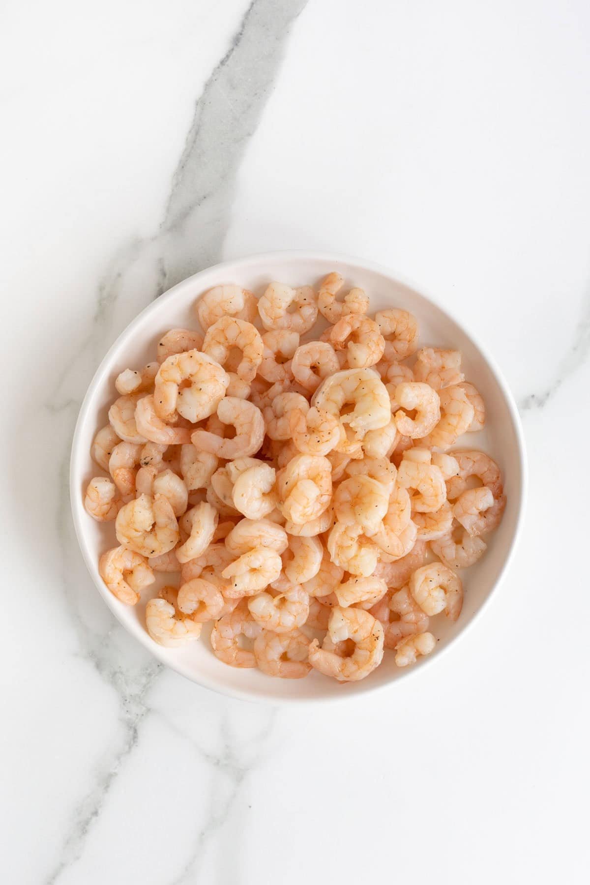 Overhead shot of a white bowl filled with cooked, pink shrimp, lightly seasoned, on a white marble surface