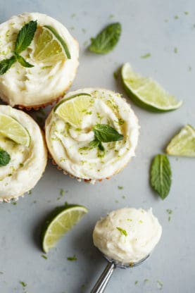Overhead shot of some Lemon Cupcakes scattered against gray background with lime slices and mint