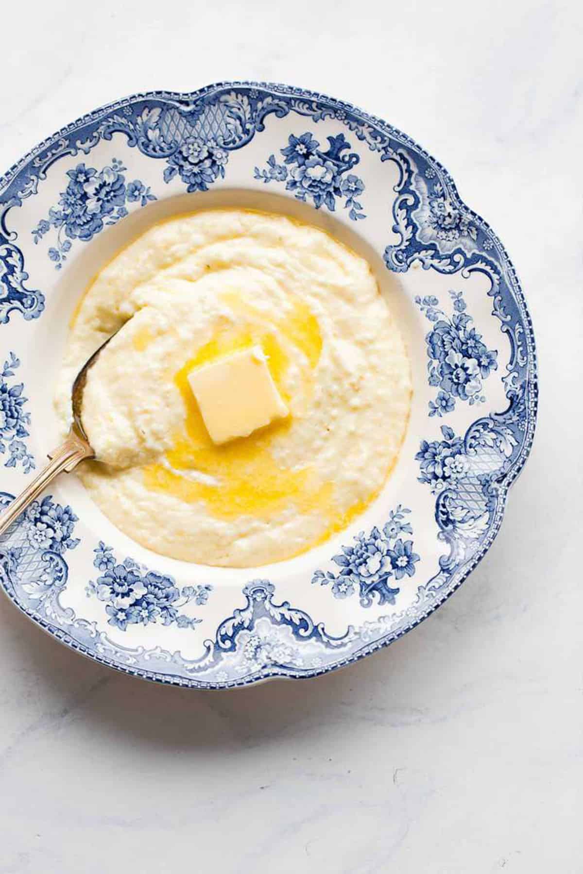 Homemade grits in a blue and white bowl on the table with a spoon in it and a pat of butter melting on top.