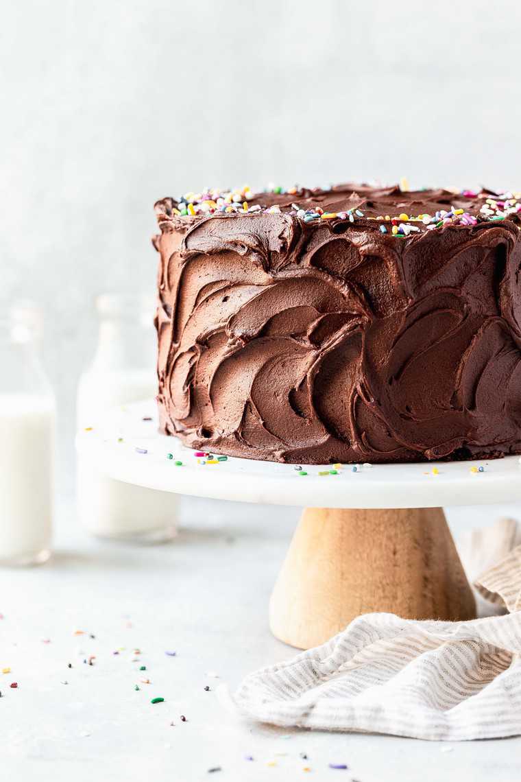 Frosted chocolate birthday cake with sprinkles on top on a cake stand against white background