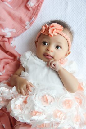 Three Months Old Baby Cakes Harmony smiling and dressed in a white and peach dress and a peach flower headband around her forehead