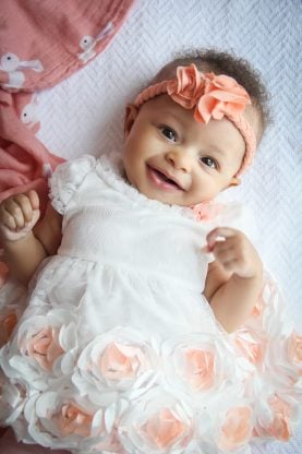 Three Months Old Baby Cakes Harmony smiling and dressed in a white and peach dress and a peach flower headband around her forehead