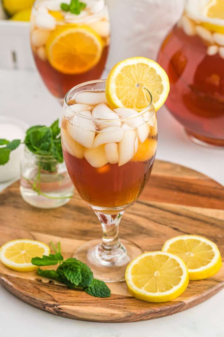 Two glasses filled with ice and sweet tea with lemon wedges on the side sitting on a wooden cutting board with mint nearby