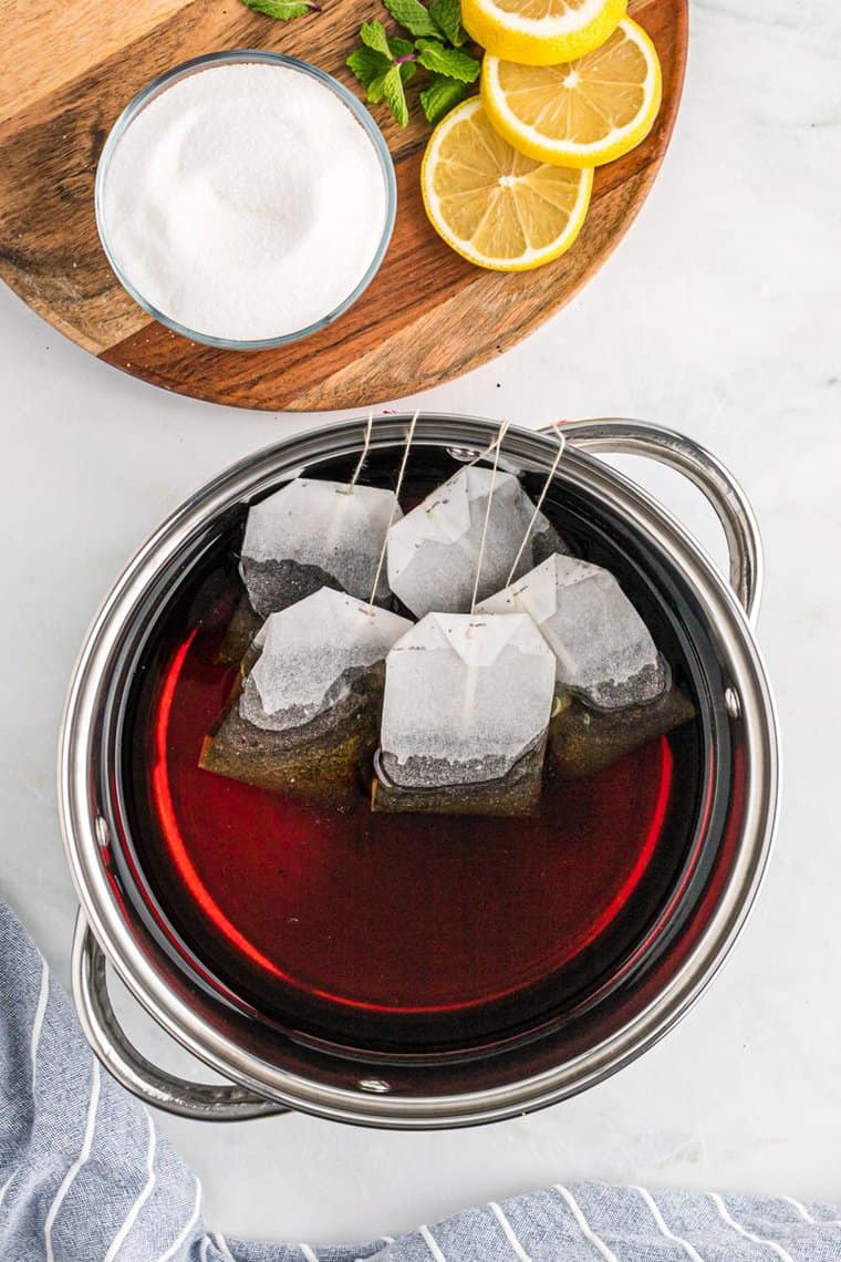 An overhead of five black tea bags steeping in a large silver pot with a bowl of sugar and lemon slice nearby