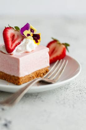 Close up of a piece of No Bake Strawberry Cheesecake served on a white plate with a fork and topped with a sliced strawberry, whipped cream and a flower