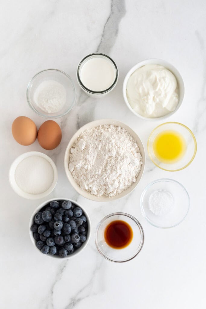 ingredients in clear and white bowls to make blueberry pancake recipe on white countertop