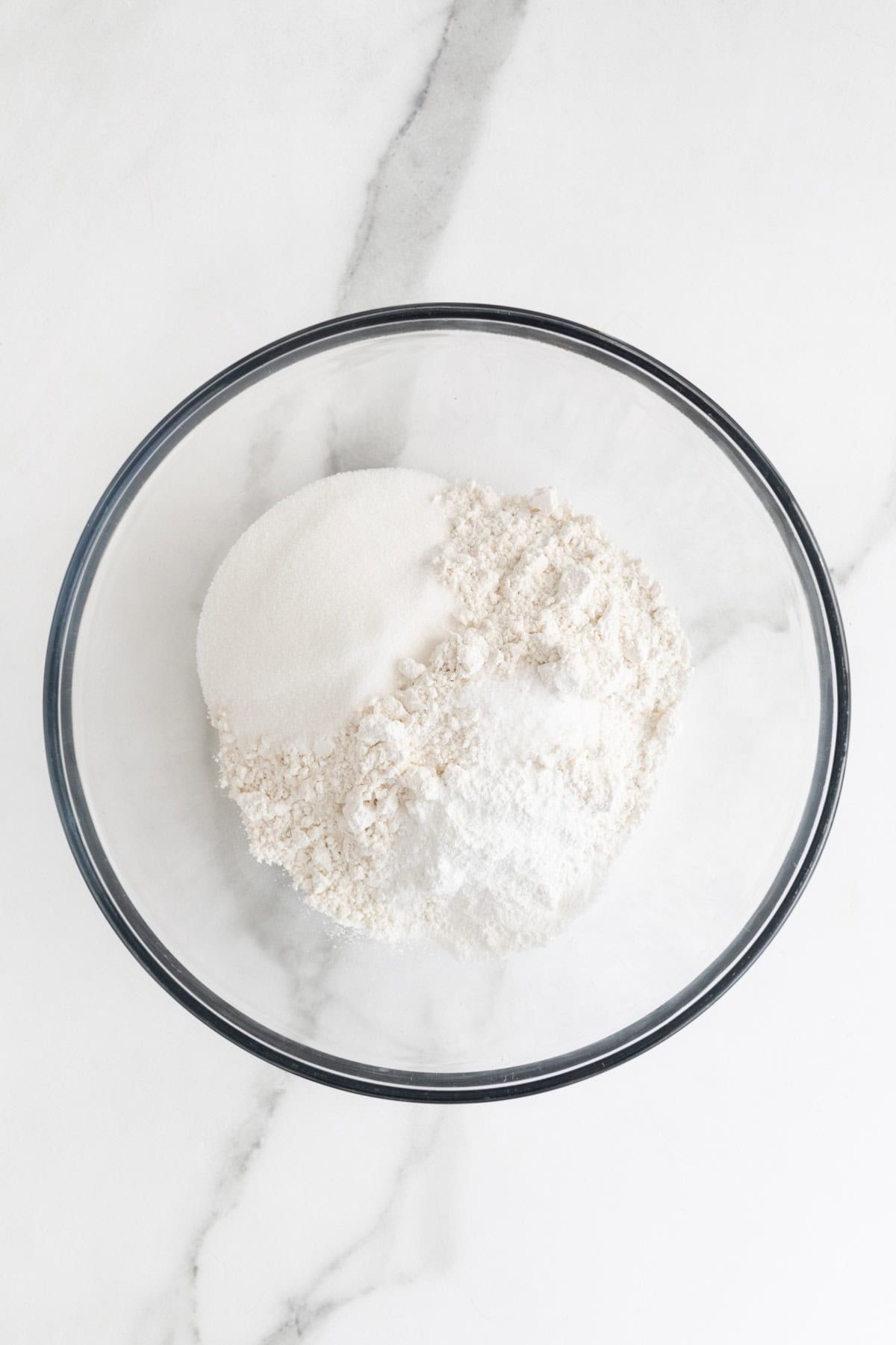 Flour, sugar and leavening added to a clear bowl on white countertop